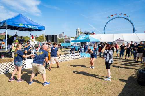 A lively outdoor event with people in referee shirts playing a game, surrounded by tents and a Ferris wheel in the background.