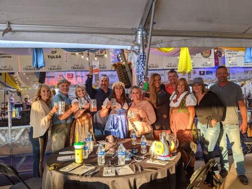 A group of people in festive attire holding drinks under a tent, with colorful decorations in the background.