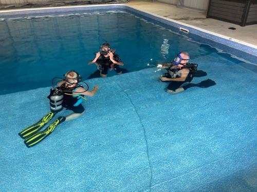 Three divers in scuba gear are underwater in a pool, engaged in conversation. Bright blue water surrounds them.