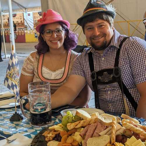 A man and woman in traditional Bavarian attire enjoy a festive table with food and drinks at an outdoor event.