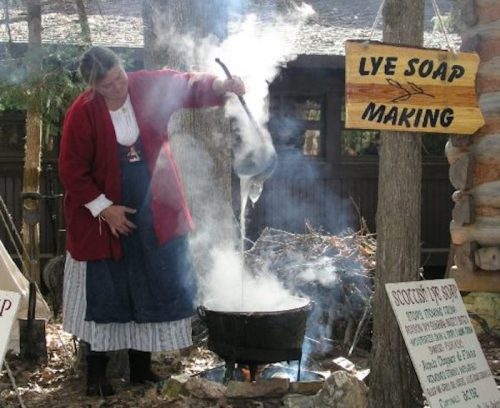 A woman in period clothing stirs a pot over a fire, with smoke rising and a sign for lye soap making nearby.