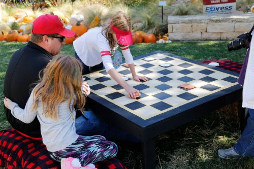 A man and two children play a large checkers game outdoors, surrounded by pumpkins and autumn scenery.