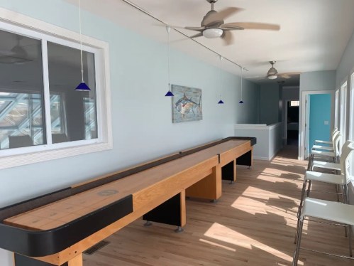 A bright room with a shuffleboard table, bar stools, and blue pendant lights, featuring light wood flooring and a coastal theme.