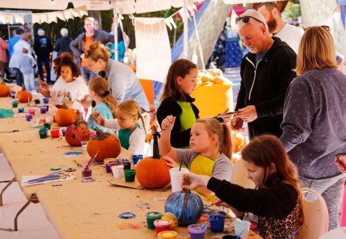 Children and adults paint pumpkins at a festive outdoor event, surrounded by colorful supplies and decorations.