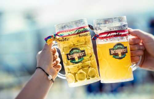 Two hands holding large beer mugs with colorful ribbons, celebrating Oktoberfest.