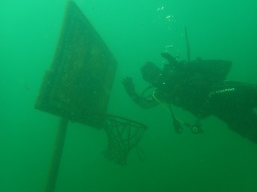 A diver explores an underwater basketball hoop in murky green water.
