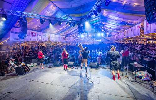 A lively concert scene with a band performing on stage in front of a large, enthusiastic crowd under a colorful tent.