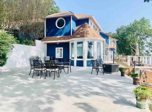 A blue house with a round window, surrounded by trees, features a patio with outdoor seating and a view of the water.
