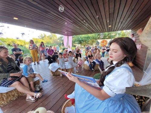 A storyteller in a blue dress reads to a captivated audience of children and adults in a cozy outdoor setting.