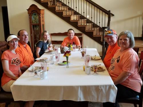A group of six people in orange shirts sitting around a dining table with food and drinks, smiling at the camera.