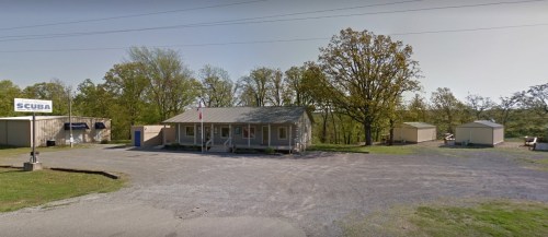 A small building with a porch, surrounded by trees and gravel, featuring a "SCUBA" sign and storage sheds nearby.