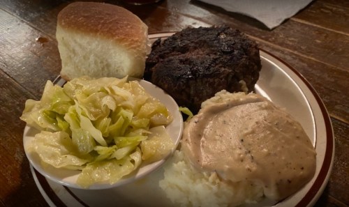 A plate with a hamburger patty, mashed potatoes with gravy, sautéed cabbage, and a roll on a wooden table.