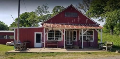 A rustic red building with a metal roof, surrounded by greenery, featuring a porch and outdoor seating.