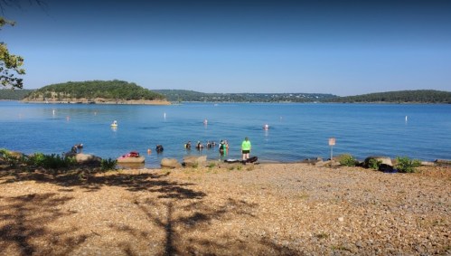 A serene lake scene with people swimming, a rocky shore, and green hills in the background under a clear blue sky.
