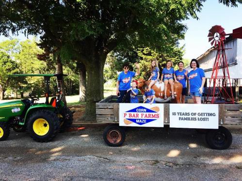 A group of people in blue shirts pose on a wagon with a tractor nearby, celebrating 10 years of P Bar Farms Maze.