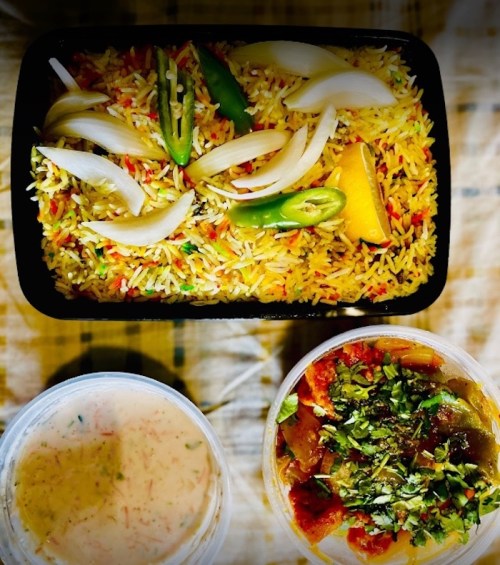 A container of colorful biryani topped with onions and green chilies, alongside two bowls of raita and curry.