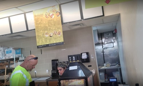 A man in a bright shirt interacts with a woman at a food counter, with a menu displayed above them.