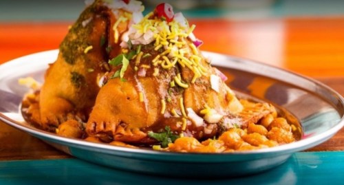 A plate of colorful chaat featuring two crispy samosas topped with chutney, onions, and sev, served with chickpeas.
