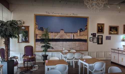 Interior of a café featuring a large mural of the Louvre museum, with tables, chairs, and decorative plants.