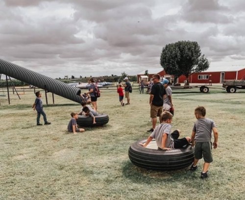 Children playing on tires at a farm, with adults supervising and a cloudy sky in the background.