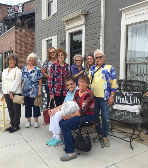 A group of women poses together outside a shop, smiling and dressed in casual attire.