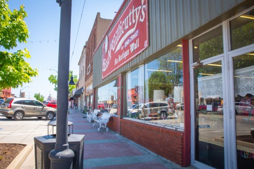 A sunny street view of a small town with shops, featuring a sign for "Antiquities" and white benches outside.