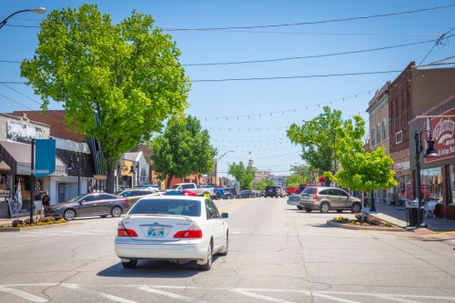 A sunny street scene with shops, trees, and parked cars, leading to a distant clock tower.