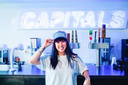 A smiling woman in a gray shirt and cap poses in front of a neon sign that reads "CAPITALS" at an ice cream shop.