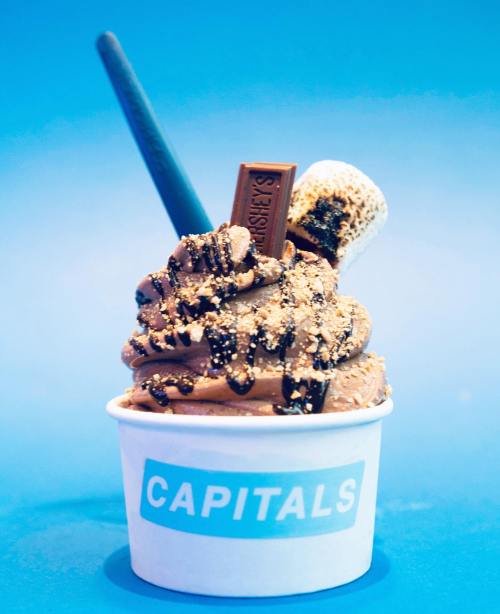 A cup of chocolate soft serve topped with a Hershey's bar, marshmallow, and chocolate drizzle against a blue background.