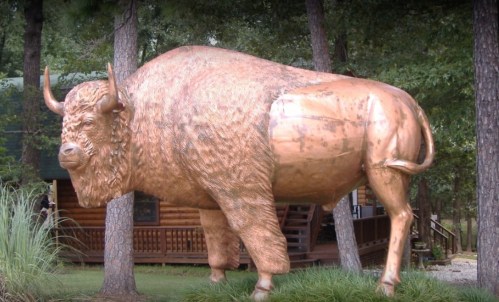 A large bronze statue of a bison stands among trees, with a rustic building visible in the background.