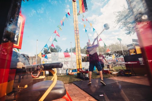 A person swings a mallet at a carnival game, with colorful flags and a tall scoring tower in the background.