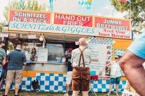 A colorful food stall at a festival, featuring signs for schnitzels, fries, and bratwurst, with people in line.