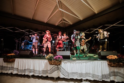 A lively band in traditional attire performs on stage, surrounded by instruments and colorful flower arrangements.