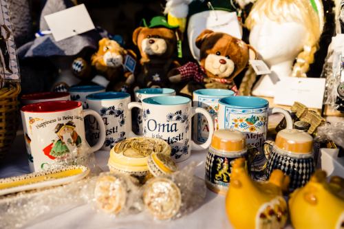 Colorful mugs with various designs and text, surrounded by stuffed animals and decorative items at a market stall.