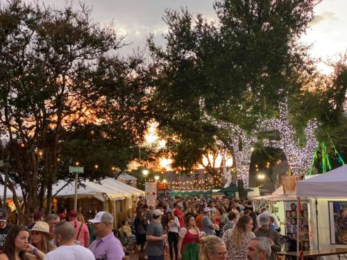 A bustling outdoor market at sunset, filled with people, tents, and festive lights among trees.