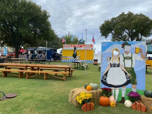 Outdoor festival scene with food stalls, picnic tables, and a colorful backdrop featuring a couple in traditional attire.