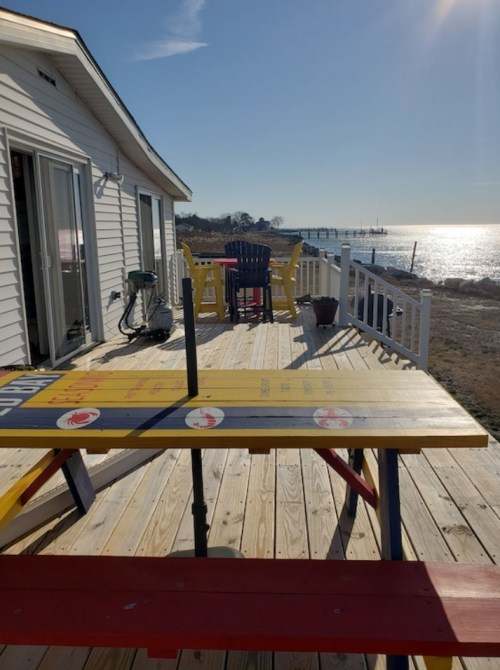 A sunny deck with colorful picnic tables overlooking a calm water view and distant pier.