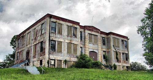 Abandoned building with peeling paint and boarded windows, surrounded by overgrown grass under a cloudy sky.
