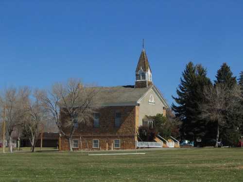 A stone building with a steeple, surrounded by trees and a clear blue sky, set in a grassy area.