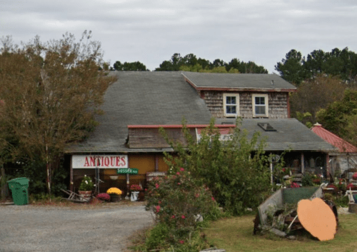 A rustic building with a sign reading "Antiques," surrounded by greenery and vintage items in a rural setting.