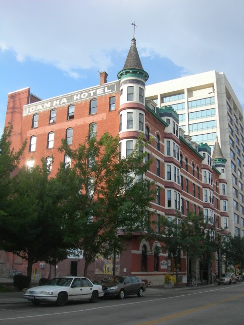 Historic Idanha Hotel with a turret, surrounded by trees and modern buildings, under a partly cloudy sky.