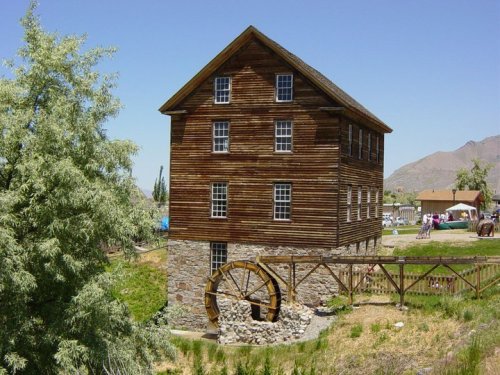 A historic wooden mill with a stone foundation and a water wheel, surrounded by trees and mountains under a clear blue sky.