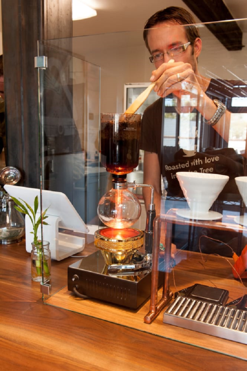 A barista brews coffee using a siphon coffee maker, stirring the mixture with a wooden stick.