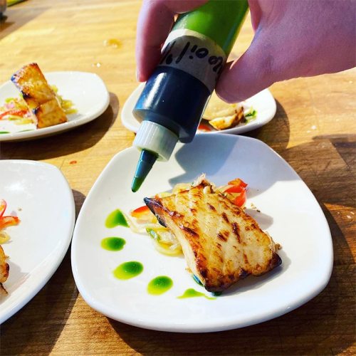 A hand drizzles green sauce over a piece of grilled fish on a white plate, garnished with colorful vegetables.
