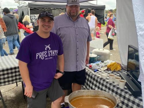 Two men stand beside a large pot of food at an outdoor event, with tents and a crowd in the background.