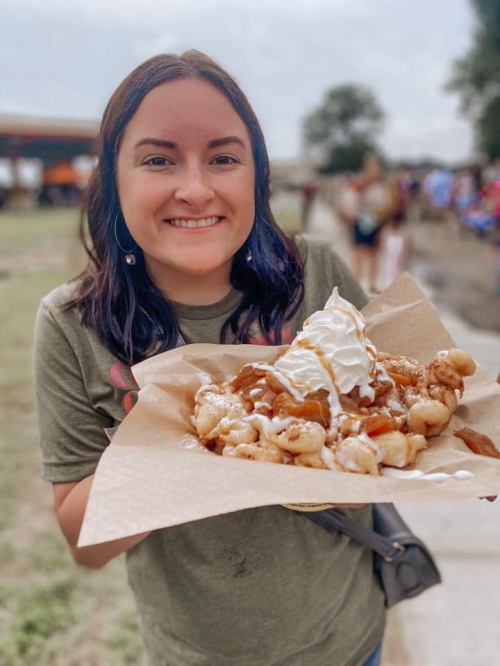 A smiling woman holds a large plate of funnel cake topped with whipped cream and syrup at an outdoor event.