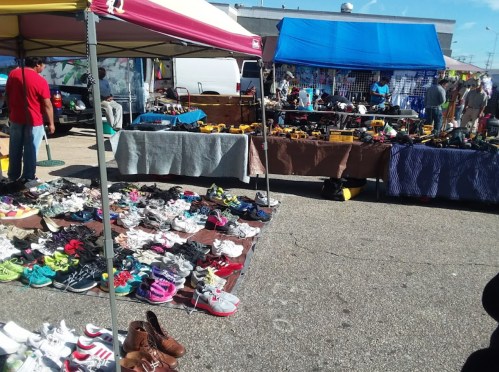 A bustling market scene with various shoes displayed on tables and the ground, under colorful tents.
