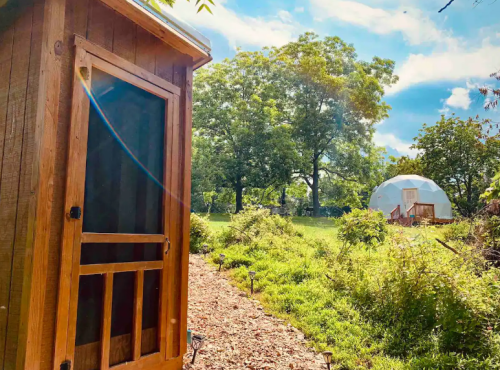 A wooden structure with a screen door beside a path, leading to a geodesic dome in a lush green landscape.