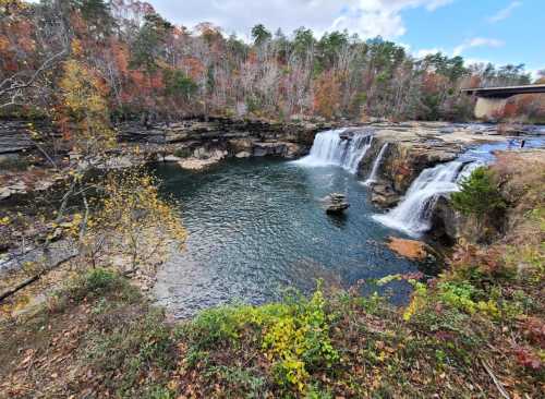 A scenic view of a waterfall cascading into a serene pool, surrounded by autumn foliage and rocky terrain.