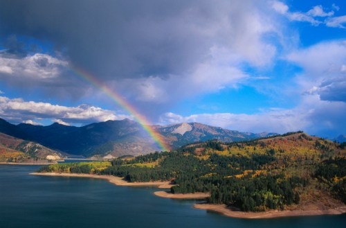 A vibrant rainbow arcs over a serene lake surrounded by mountains and lush greenery under a partly cloudy sky.